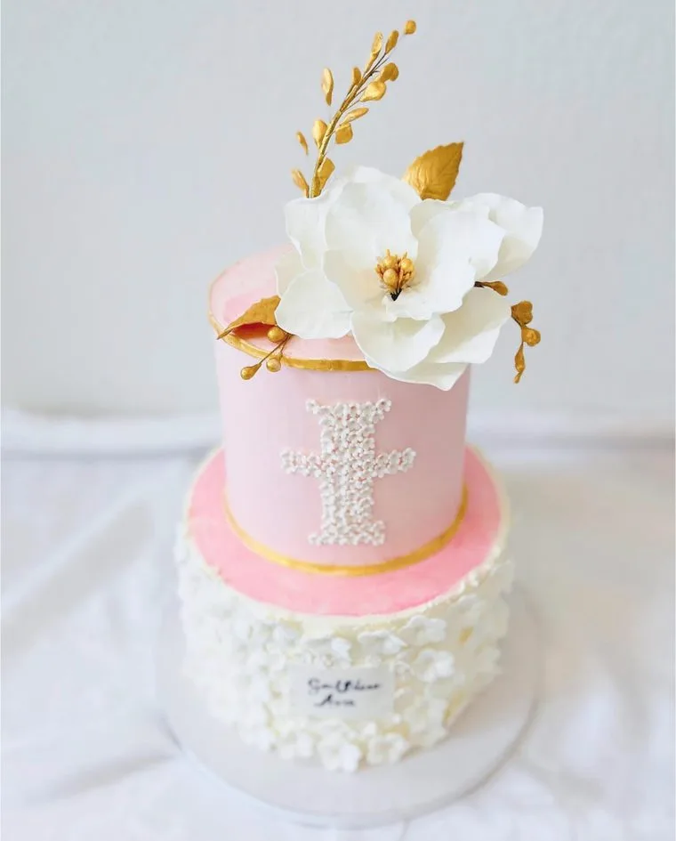 Two-tier pink and white cake with a sugar magnolia, gold leaves and a piped cross